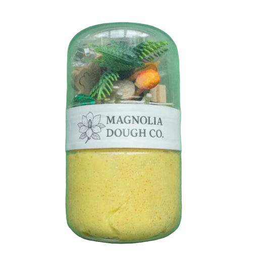 Jar of yellow dough with decorative elements on a green background, featuring Magnolia Dough Co. label.