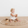 Baby sitting on a round beige mat with wooden toys on a light wood floor.