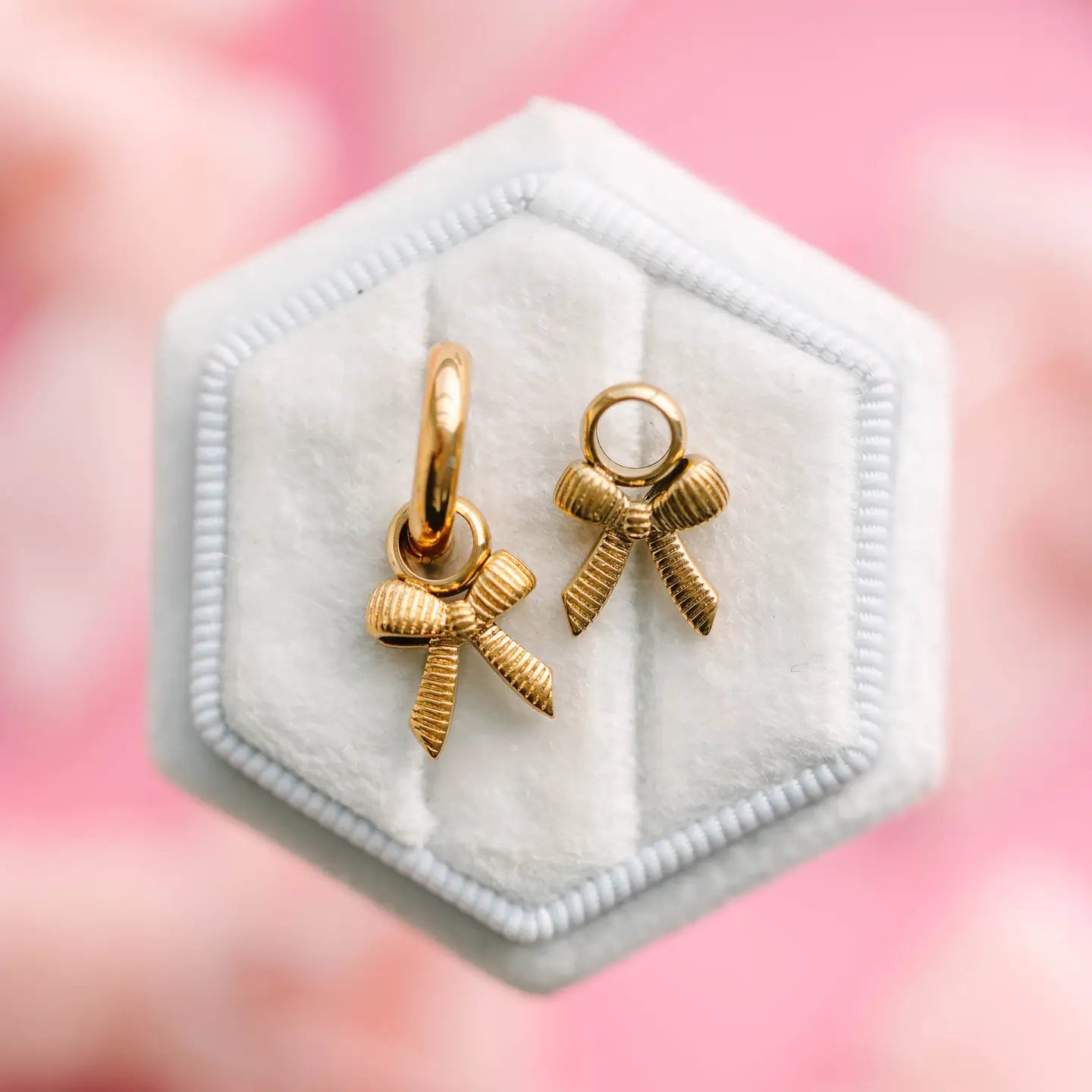 Gold bow-shaped earrings in a hexagonal white jewelry box with a pink blurred background