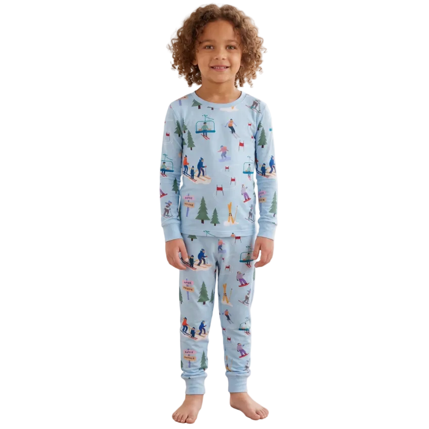A young child with curly hair stands barefoot in a bright bedroom, wearing a light blue long-sleeve pajama set with a colorful allover winter sports and outdoor print. The room has a bed with cream bedding, a window with sheer curtains, and a small shelf with decorative items.