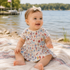 Baby sitting on a blanket by a lake wearing a dress with a Fourth of July theme graphic