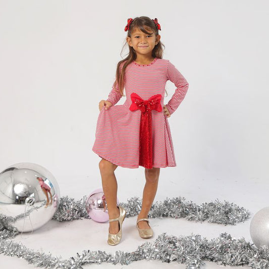 Young girl in a red and white striped dress with a large bow standing in front of Christmas decorations.