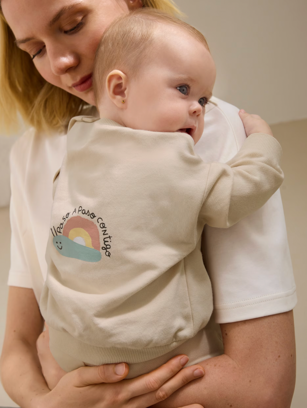 Woman holding a baby wearing a beige sweater with a rainbow design.