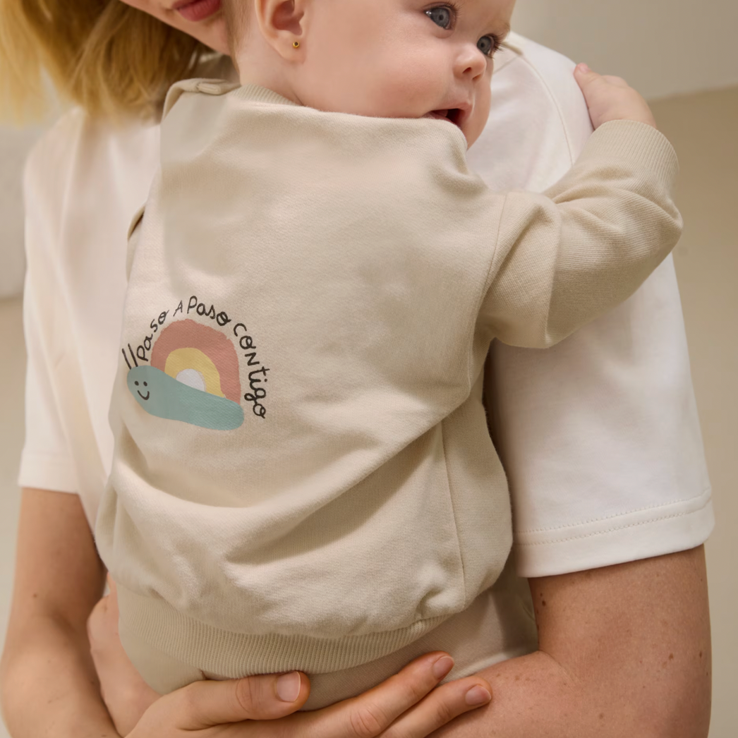 Woman holding a baby wearing a beige sweater with a rainbow design.