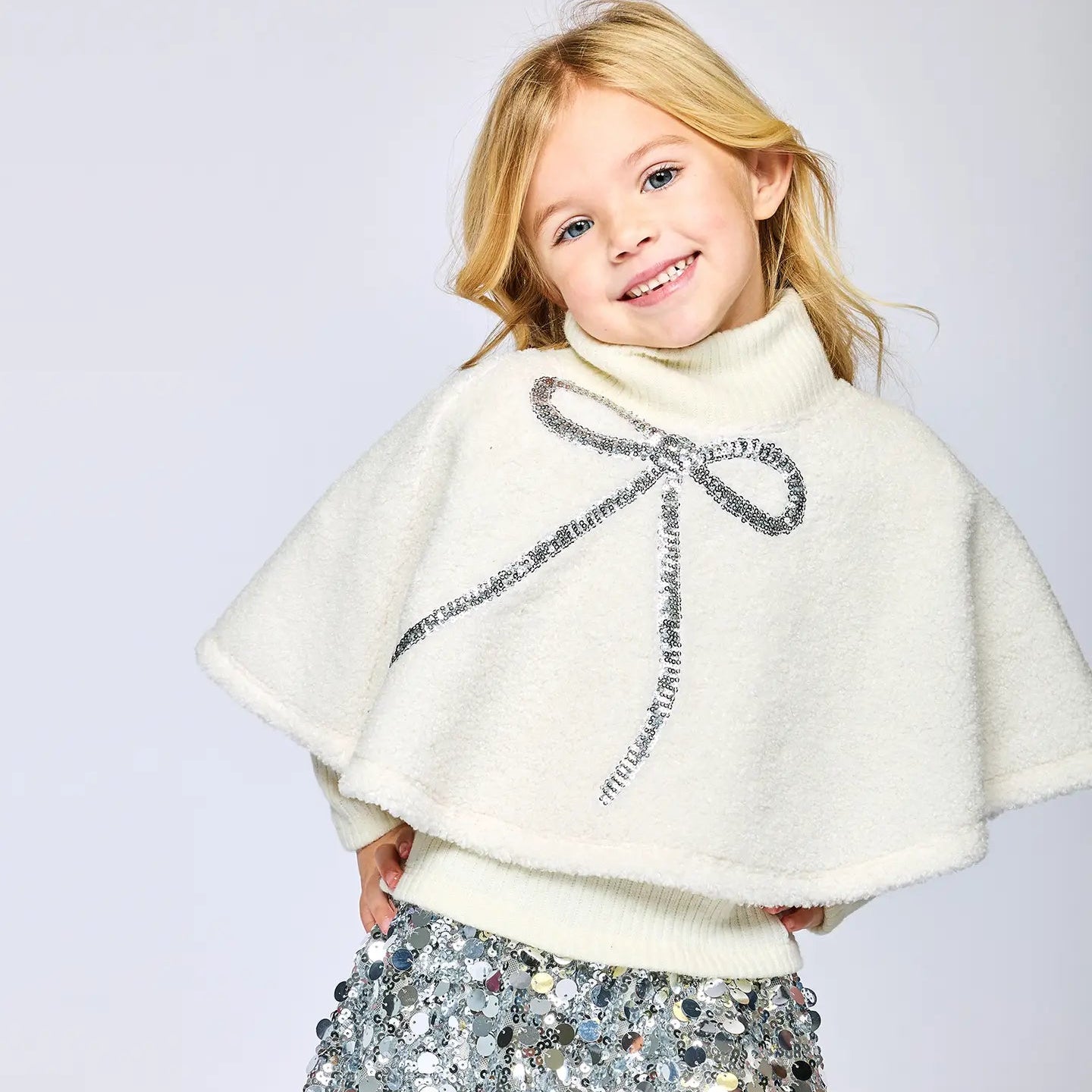 Child wearing a white sherpa poncho with a sequin bow and a silver sequin skirt on a light gray background