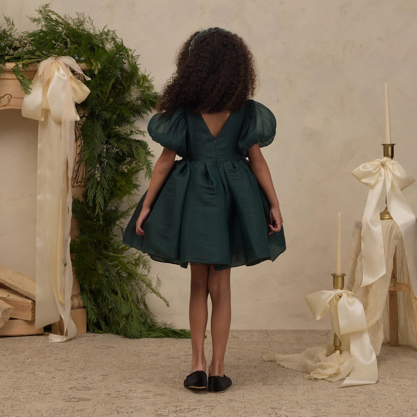 A young girl is shown from behind wearing a dark green puff-sleeve dress with a tie-back back.
