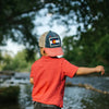 Child wearing a Colorado flag cap and red shirt standing by a river with trees in the background