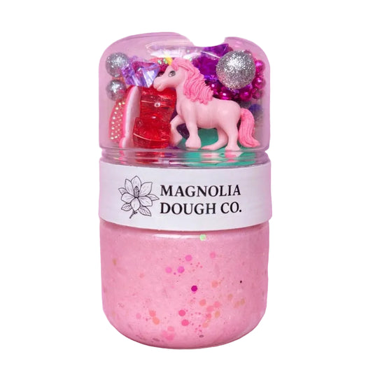 Pink dough with unicorn and glittery decorations in a container labeled 'Magnolia Dough Co.' on a pink background.