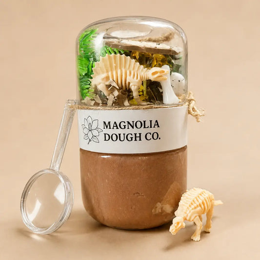 A clear dome-topped jar filled with miniature paleontology toys, including a dinosaur skeleton and a small white animal figure, wrapped with a white label reading 'MAGNOLIA DOUGH CO.' and set on a beige background with a magnifying glass and small dinosaur toy nearby.