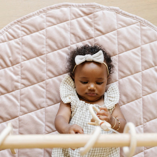 Baby lying on a quilted pink surface wearing a white bow headband and checkered outfit.
