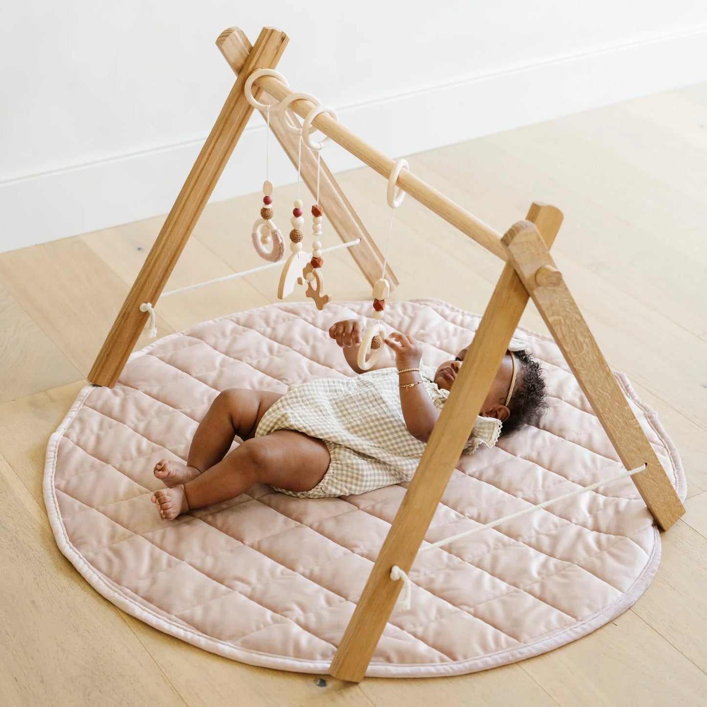 Baby lying on a quilted play mat with a wooden teepee structure and hanging toys.