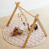 Baby lying on a quilted play mat with a wooden teepee structure and hanging toys.