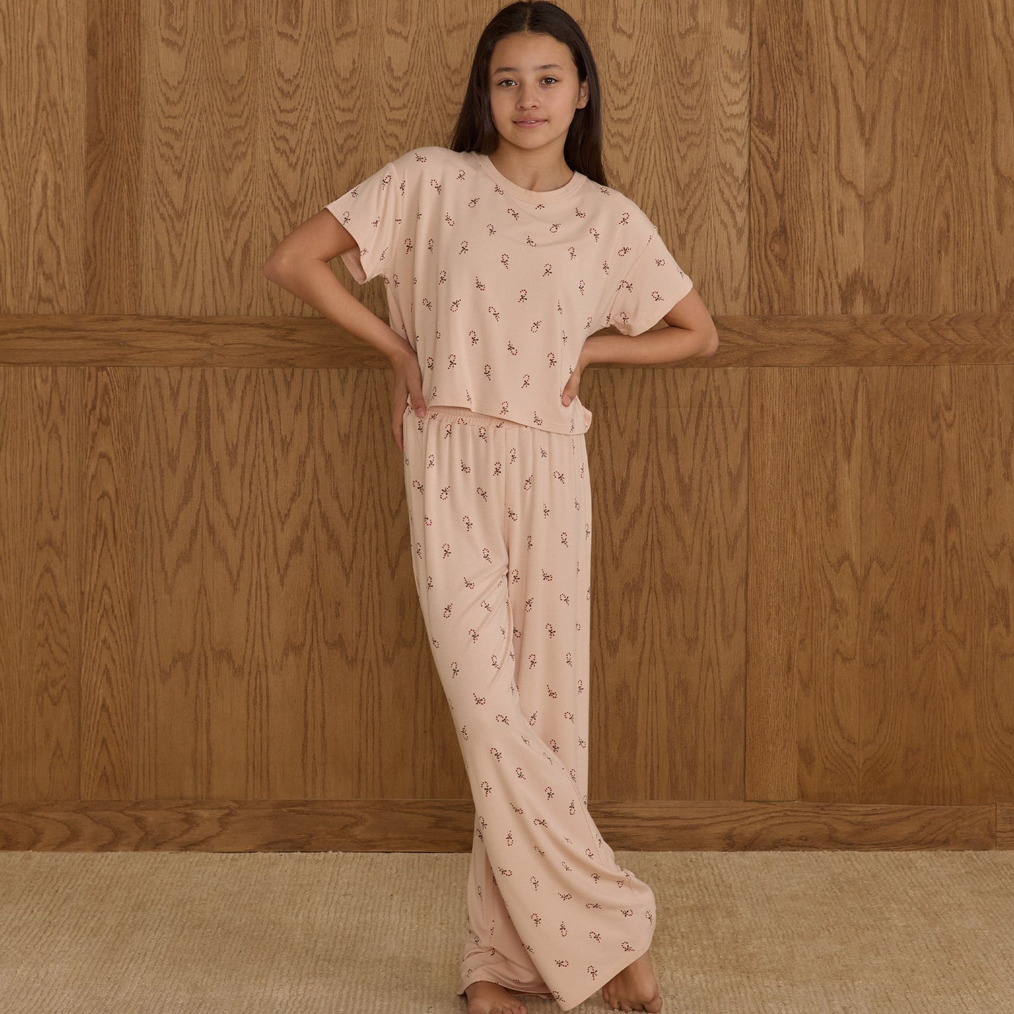 Tween wearing a light pink outfit with a subtle pattern against a wooden paneled wall.