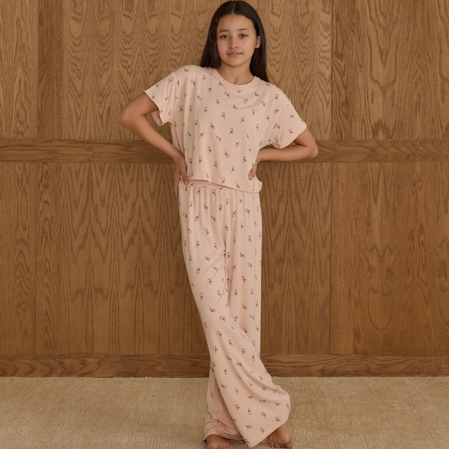 Tween wearing a light pink outfit with a subtle pattern against a wooden paneled wall.