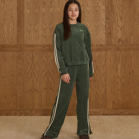 Person wearing a green tracksuit with white stripes standing against a wooden paneled wall.