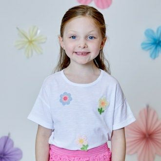 Young girl wearing a white t-shirt with floral designs and pink shorts against a decorative background.