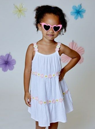 Young girl wearing a white dress with floral details and pink sunglasses against a floral background.