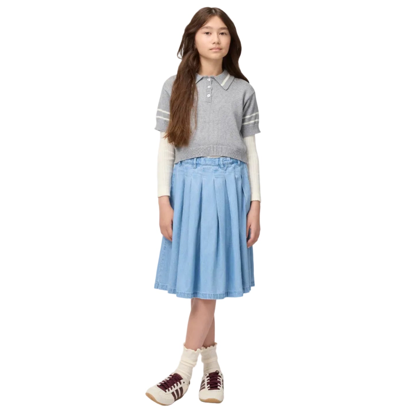 Young girl wearing a gray striped polo shirt, light blue skirt, and white sneakers on a white background
