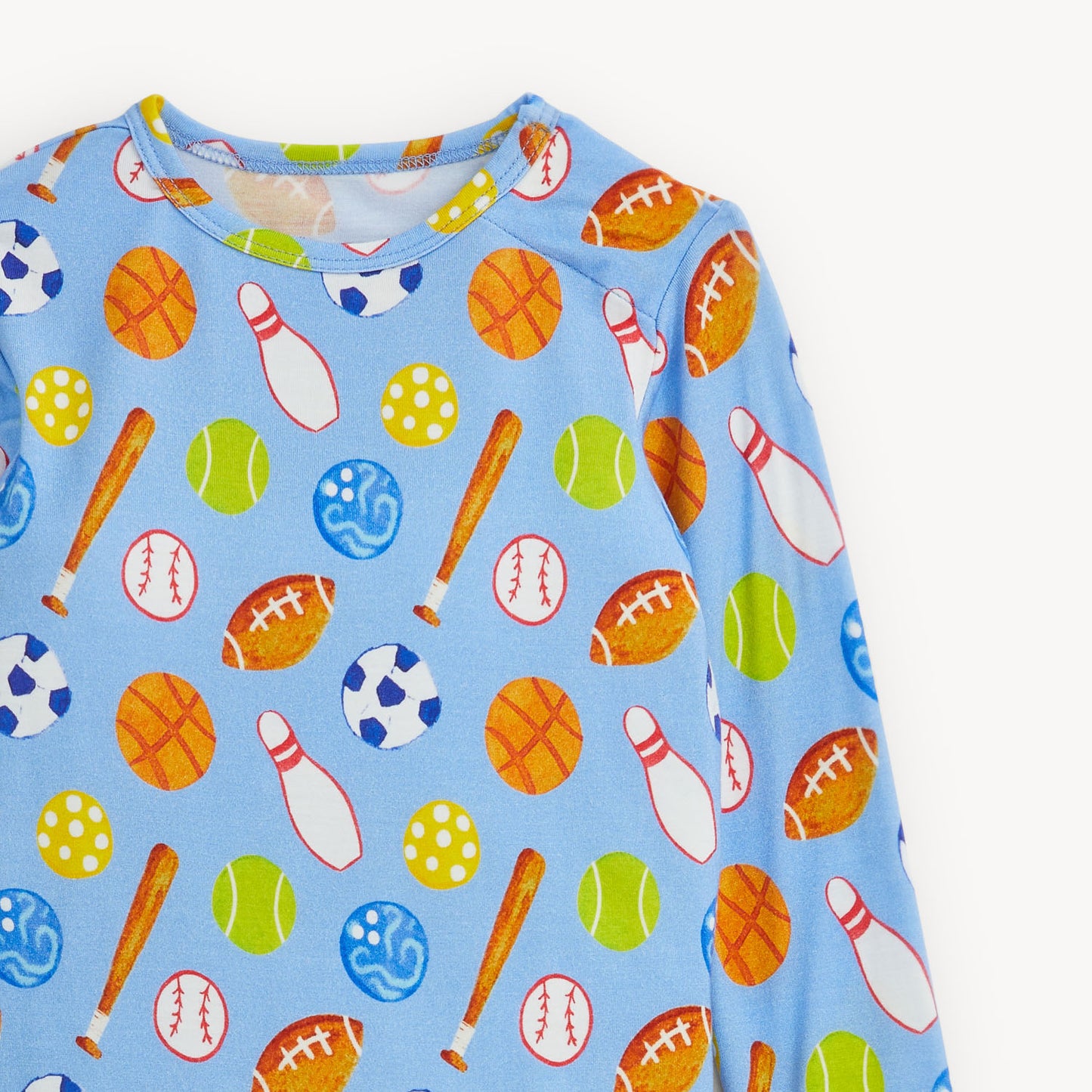 A close-up of a light blue long-sleeve top with an all-over sports-themed print featuring orange basketballs, white bowling pins, green tennis balls, brown baseball bats, and multicolored balls and bats.