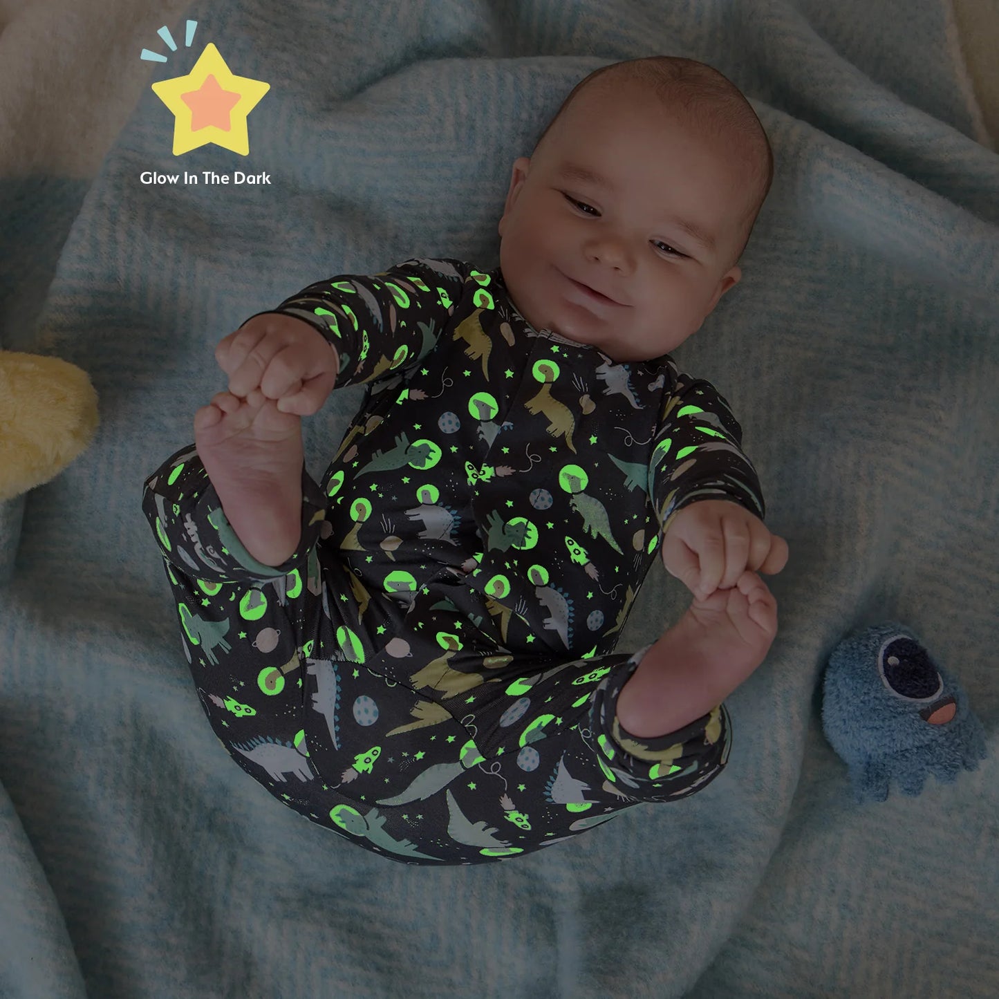 A baby lying on a textured blue blanket, wearing a long-sleeve glow-in-the-dark coverall with a green and gray dinosaur pattern on a black background.