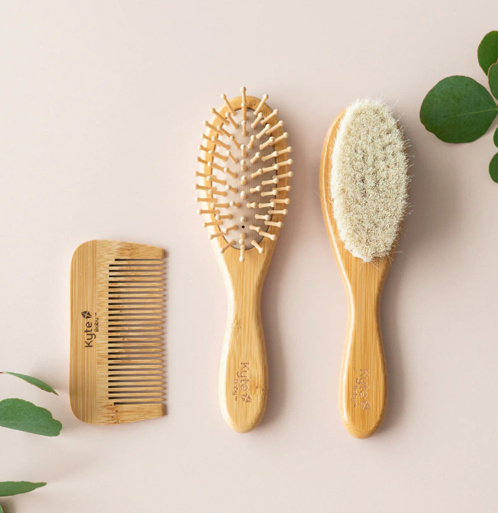 A set of three baby care brushes with bamboo handles, including a hairbrush with natural goat hair bristles, a hairbrush with wooden bristles, and a bamboo baby comb.