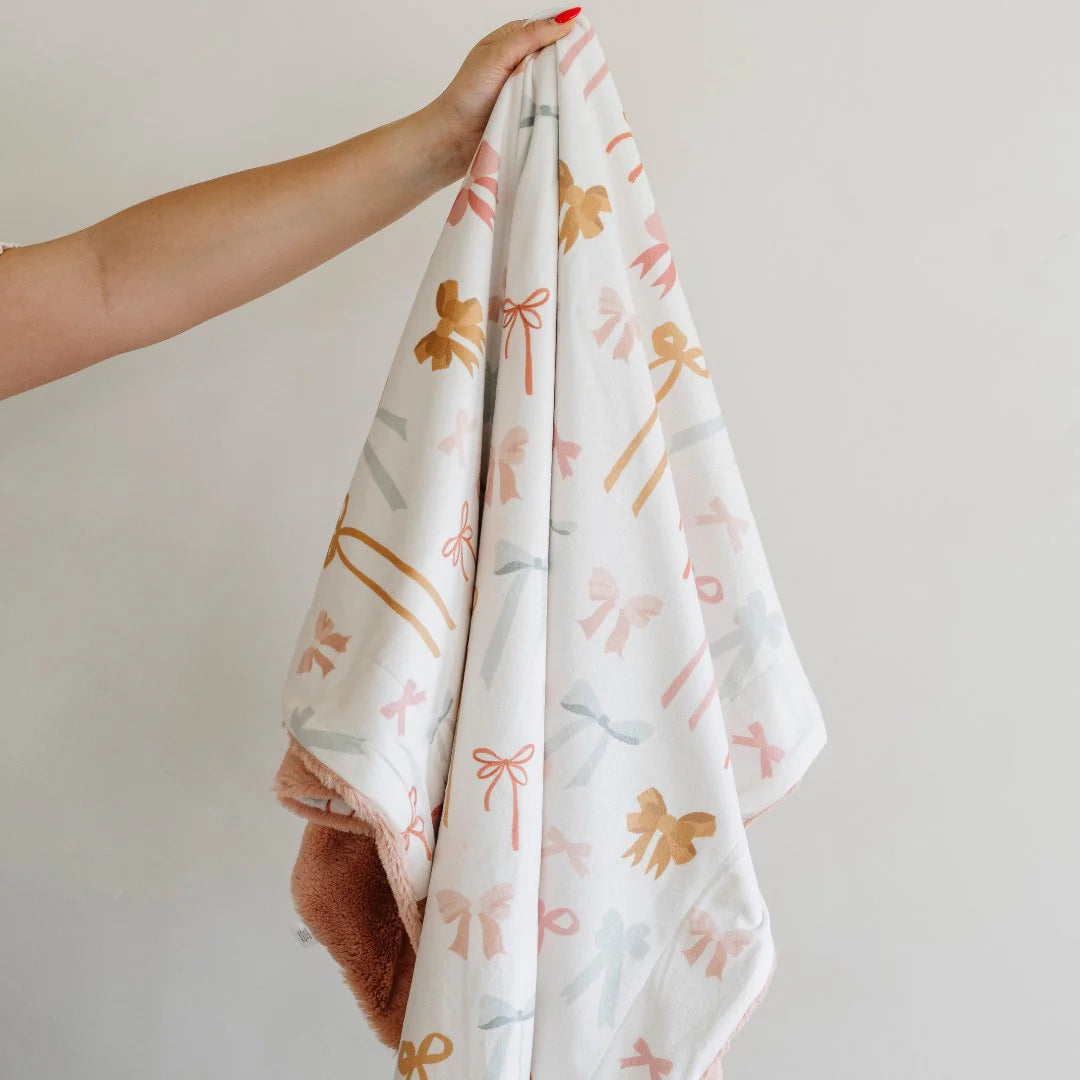 A receiving blanket with a white background featuring a pattern of minky bows on one side, and a lush pink fabric on the other side