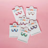 A collection of colorful huggies earrings displayed on white cards with the brand name 'Pierced Co' on them, set against a pink background.