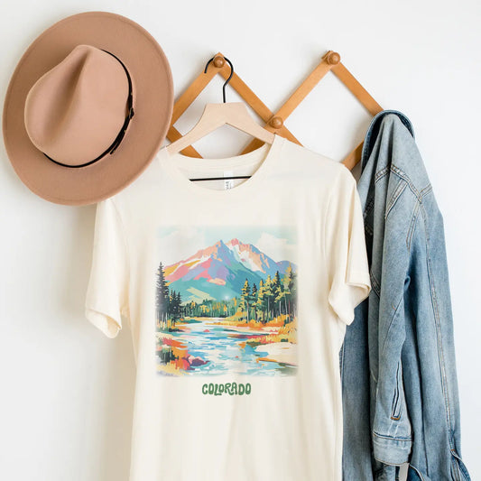White t-shirt with mountain and lake graphic, beige hat, and denim jacket on a wooden rack.
