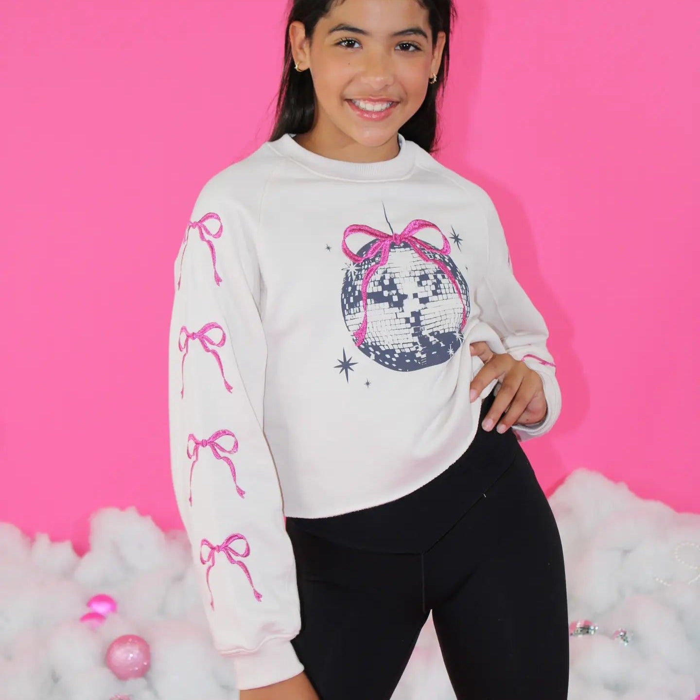 Girl wearing a white sweatshirt with pink bow designs and a disco ball graphic against a pink background.