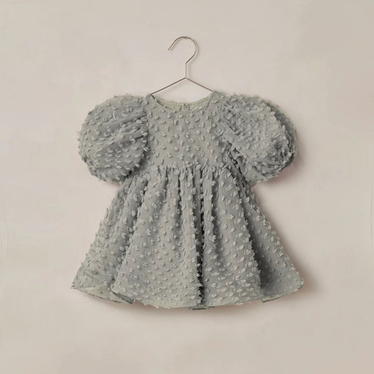 A light gray textured baby dress with puffy sleeves, hanging on a metal hanger against a plain beige wall.