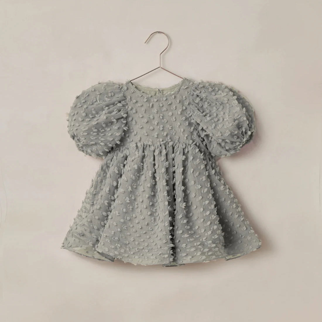 A light gray textured baby dress with puffy sleeves, hanging on a metal hanger against a plain beige wall.