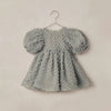 A light gray textured baby dress with puffy sleeves, hanging on a metal hanger against a plain beige wall.