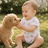 Baby in a white onesie with puppy all over print, who is sitting on grass, interacting with a small brown dog.