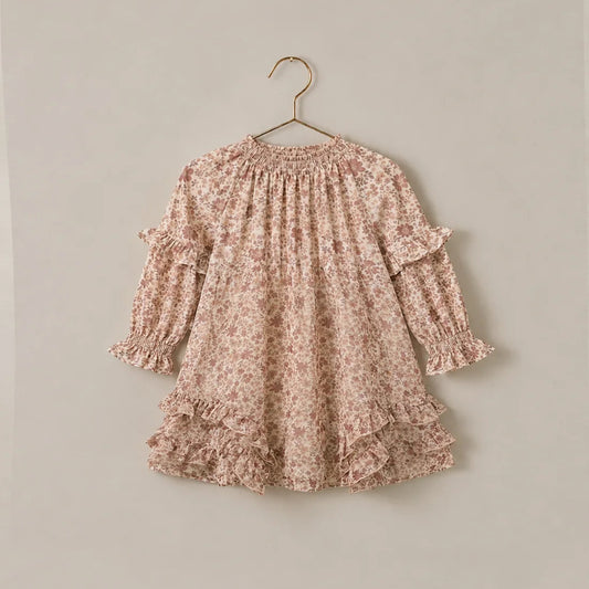 A long-sleeve babydoll dress with a delicate pink and beige floral pattern, featuring ruffled trim at the shoulders and hem, hanging on a gold hanger against a solid light beige wall.