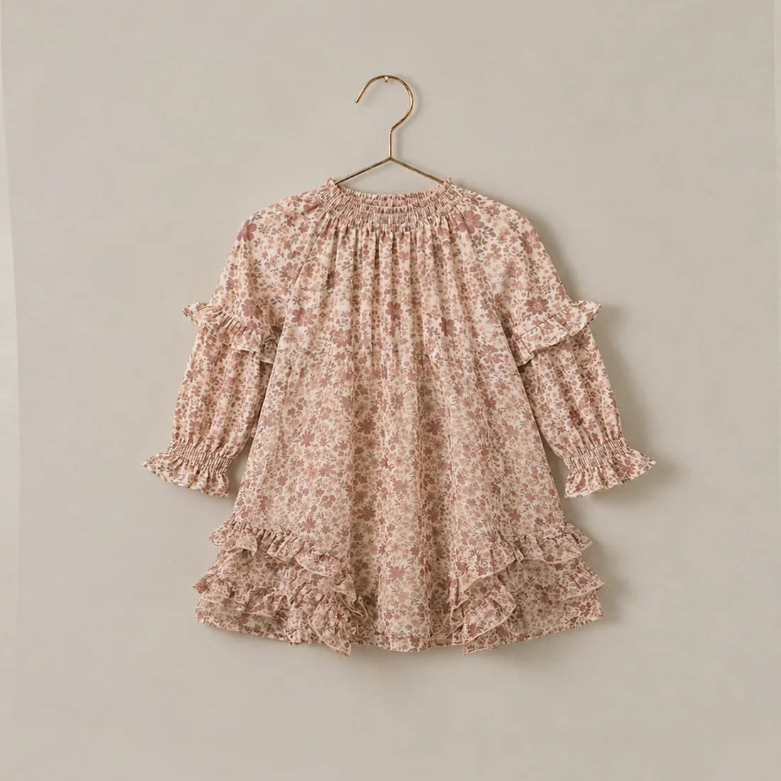 A long-sleeve babydoll dress with a delicate pink and beige floral pattern, featuring ruffled trim at the shoulders and hem, hanging on a gold hanger against a solid light beige wall.