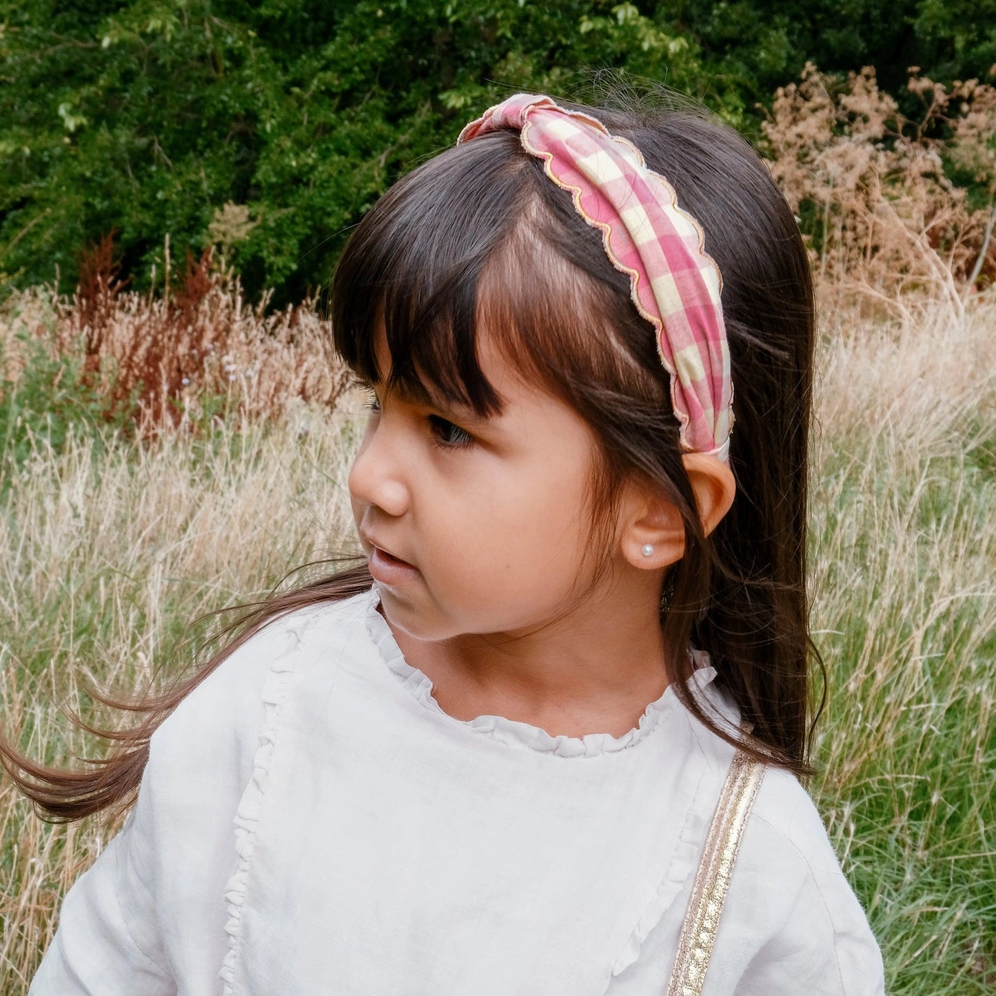 Young girl with a pink headband standing in a natural setting with greenery and dry grass.