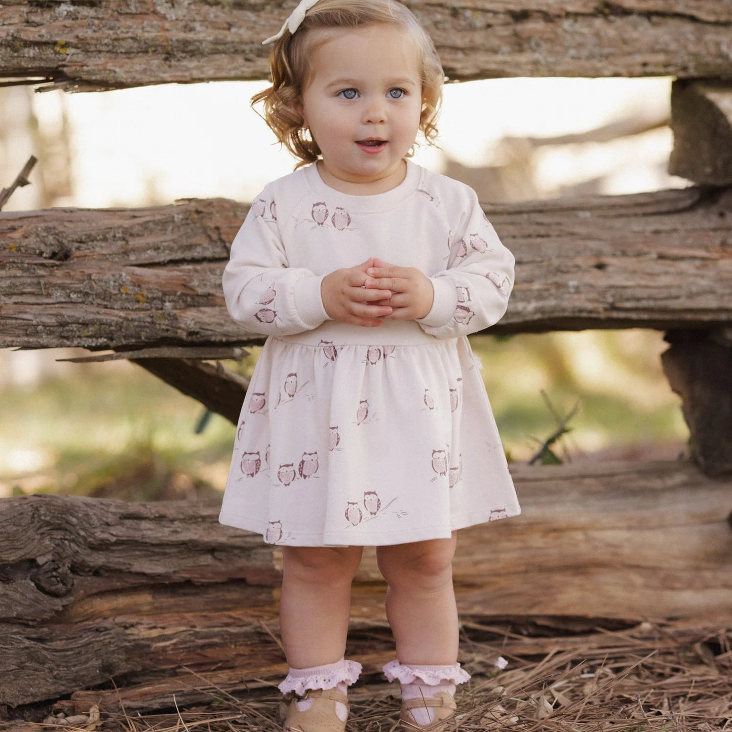A toddler girl stands outdoors in front of rustic wooden logs, wearing a long-sleeve white dress with a small pink owl print, pink ruffled boot covers, and a white bow in her hair.