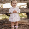 A toddler girl stands outdoors in front of rustic wooden logs, wearing a long-sleeve white dress with a small pink owl print, pink ruffled boot covers, and a white bow in her hair.
