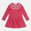A pink long-sleeve baby dress with a gathered peplum waist and a decorative embroidered yoke featuring white and green floral details, shown on a light gray background.
