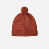 A rust-colored knit pom-pom beanie with tiny white polka dots, shown flat against a light gray background.