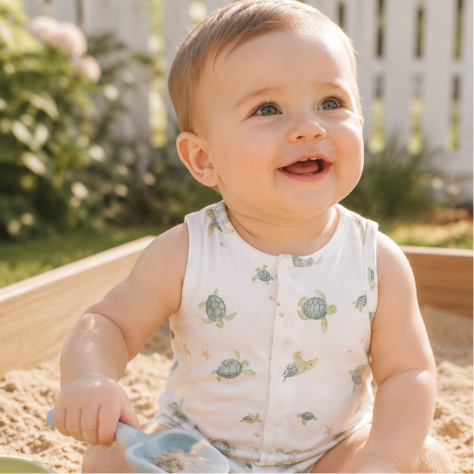 Baby playing in a sandbox with sand toys, where a white romper with green sea turtle print