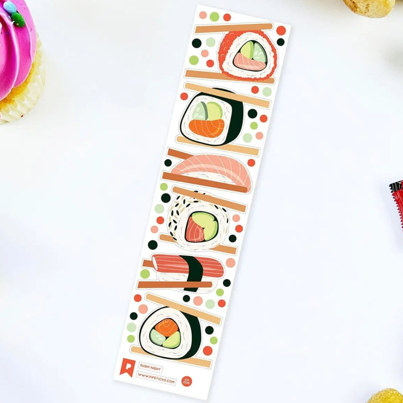A long rectangular sticker sheet featuring illustrated sushi pieces on a white background with colorful dots and chopsticks. The sheet is photographed from above on a white surface with small food props around the edges, including a pink cupcake and banana pieces.