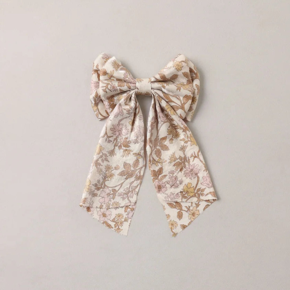A fabric hair bow with a delicate floral pattern in soft beige, brown, and muted pink, tied in a large bow shape against a plain light gray background.