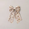 A fabric hair bow with a delicate floral pattern in soft beige, brown, and muted pink, tied in a large bow shape against a plain light gray background.