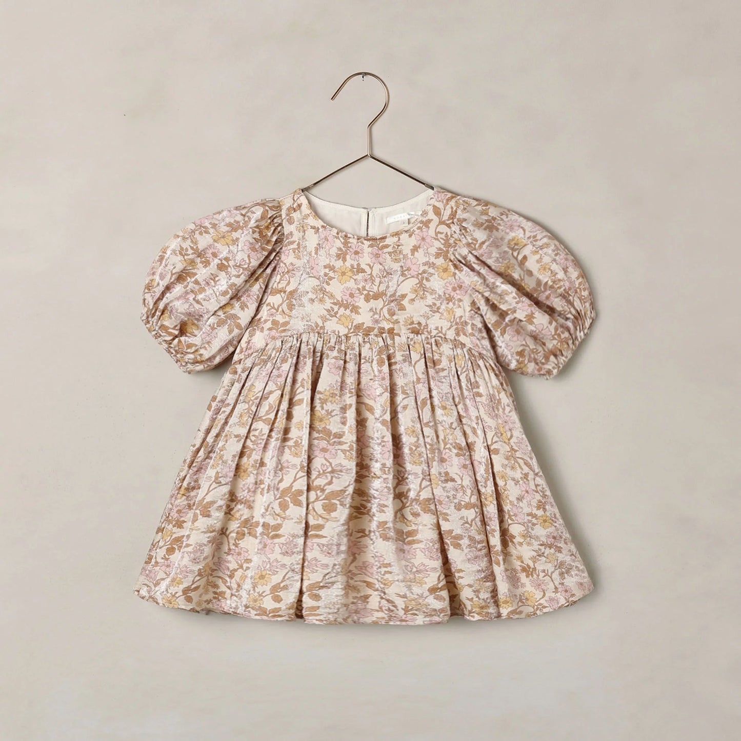 A toddler girl's dress with puff sleeves and a gathered smocked bodice, featuring an all-over floral toile pattern in beige and muted brown tones, hanging on a metal hanger against a textured beige wall.