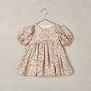 A toddler girl's dress with puff sleeves and a gathered smocked bodice, featuring an all-over floral toile pattern in beige and muted brown tones, hanging on a metal hanger against a textured beige wall.