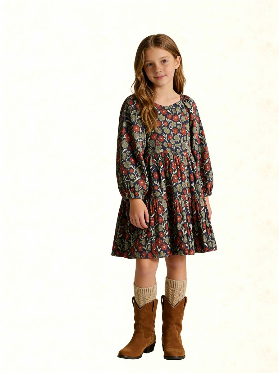 A young girl stands against a plain off-white background wearing a long-sleeve smocked dress with a dense floral and botanical print in red, green, and beige tones, paired with cream knit socks and brown boots.