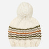 An off-white chunky knit pom-pom beanie with horizontal zigzag stripes in olive green, brown, and orange, shown against a light gray background.