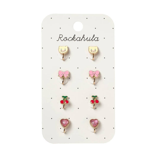 A set of six small stud earrings displayed on a white card labeled 'Rockahula'. The earrings include two yellow cat-face studs, two pink bow studs, two red cherry studs, and two pink heart studs, all with gold-tone metal settings.