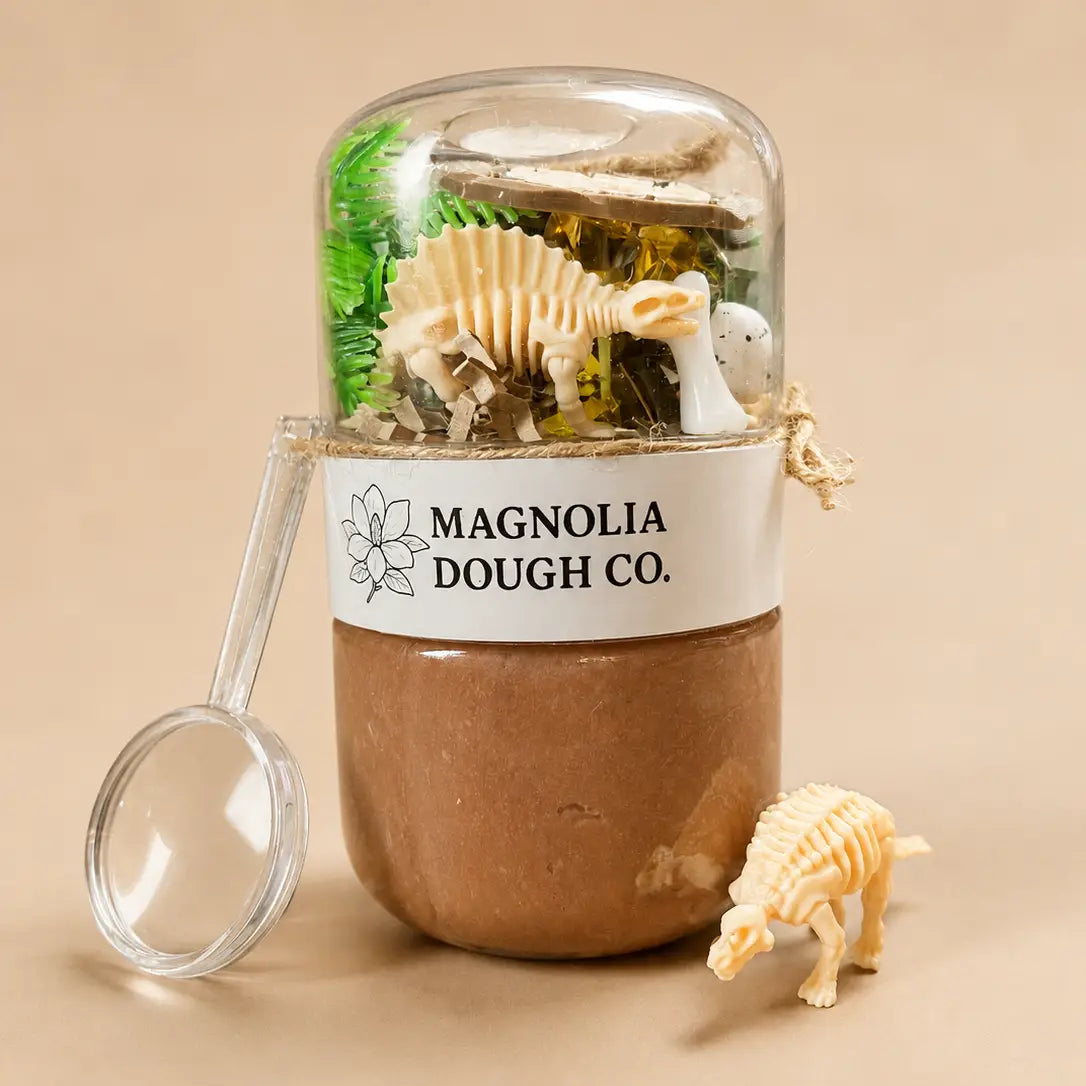 A clear dome-topped jar filled with miniature paleontology toys, including a dinosaur skeleton and a small white animal figure, wrapped with a white label reading 'MAGNOLIA DOUGH CO.' and set on a beige background with a magnifying glass and small dinosaur toy nearby.
